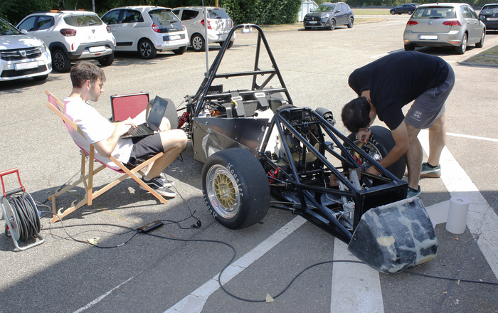 2025-07-31 PM BFFT startet erstmals in Formula Student Blick von ron auf den Rennwagen: Ein junger Mann beugt sich über den linken Vorderreifen des Rennwagens und schraubt daran, ein anderer sitzt rechts neben dem Rennwagen in einem Liegestuhl und hat ein Laptop auf dem Schoß überall liegt Werkzeug herum