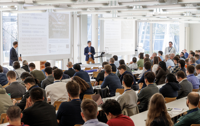 2025-04-10 Web-Meldung Modval 2025 Konferenz Zwei Männer stehen vor zahlreichen sitzenden Konferenzteilnehmenden